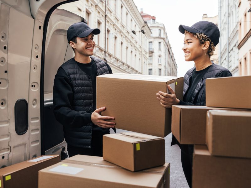 Two coworkers looking at each other while loads big cardboard box into a delivery van